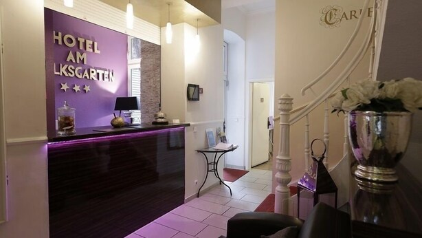 A hotel reception area with a purple sign, a reception desk, a small table with a lamp, and a decorative vase with flowers. - Hotel Am Volksgarten (Düsseldorf)
