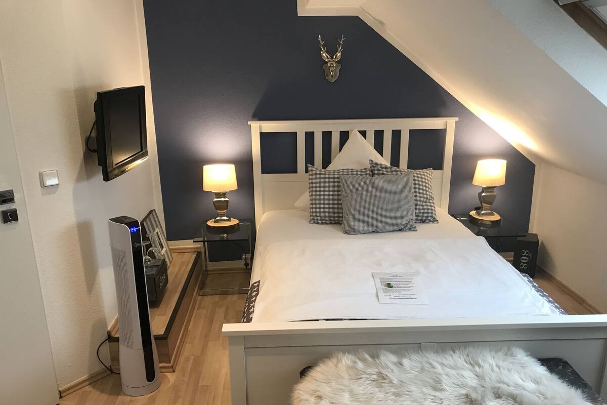 A bedroom with a white bed, two bedside lamps, a mounted TV, a small table, and a fur throw.