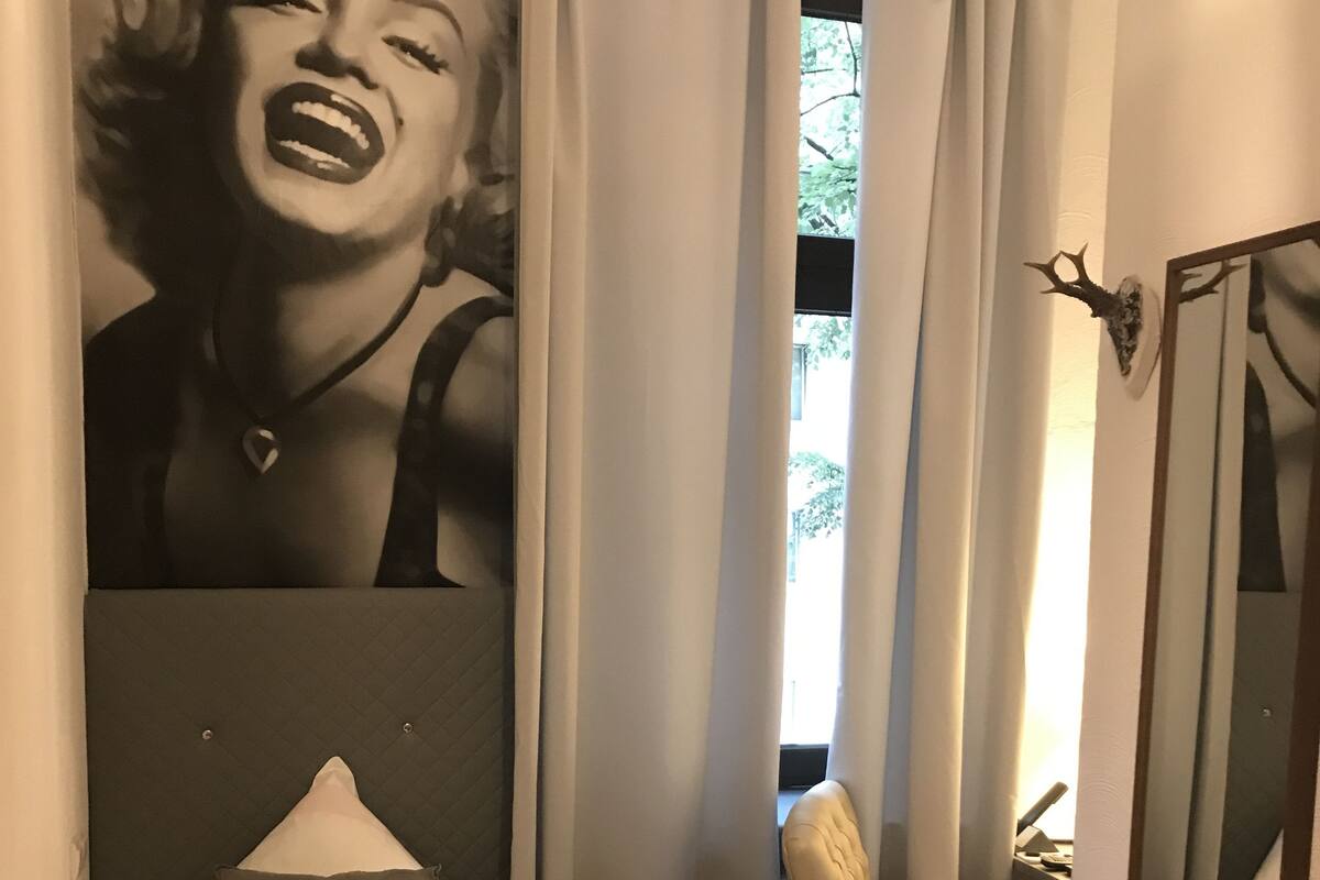 A hotel room with a bed, a small round table, a chair, a large mirror, and a wall with a black and white mural.