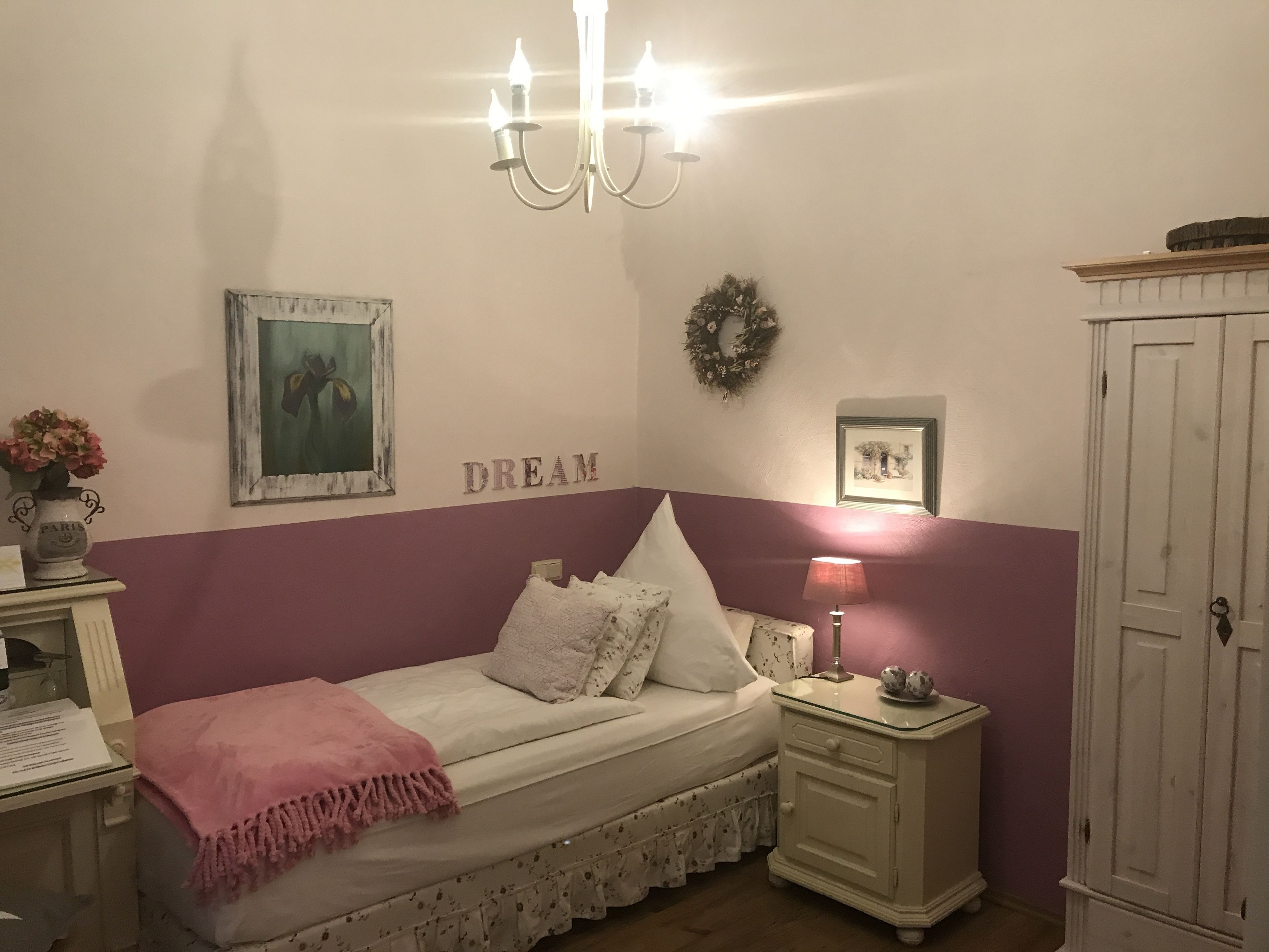 A bedroom with a bed, a nightstand, a dresser, a chandelier, and wall decorations including a painting and a wreath.