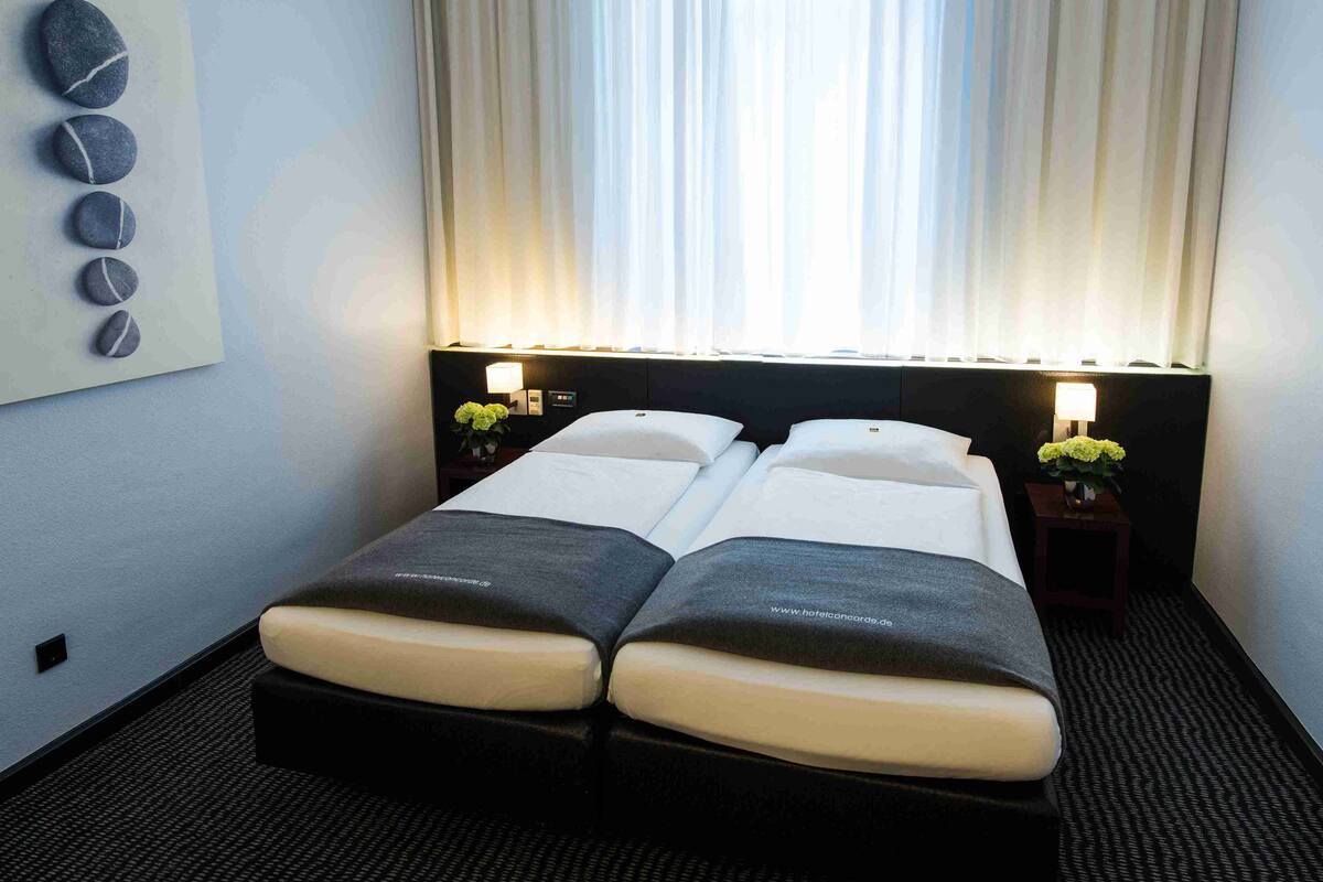 A hotel room with two beds, a nightstand with a lamp, a wall-mounted artwork, and a window with curtains.