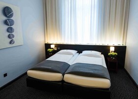Habitación de hotel con dos camas, una mesita de noche con lámpara, un cuadro en la pared y un ventanal con cortinas.