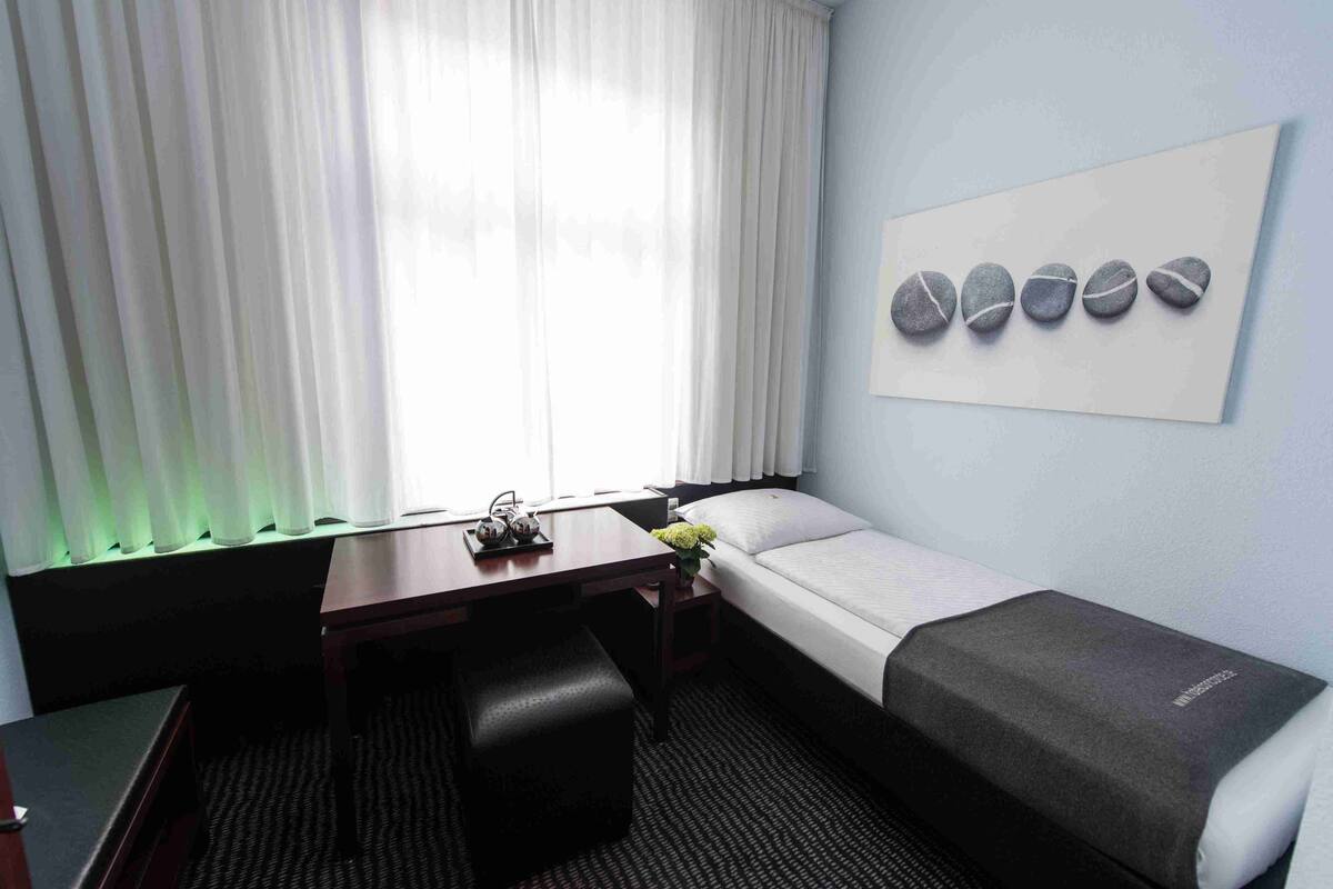 A hotel room with a bed, a desk, a chair, a small table with a vase of flowers, and a wall art piece.