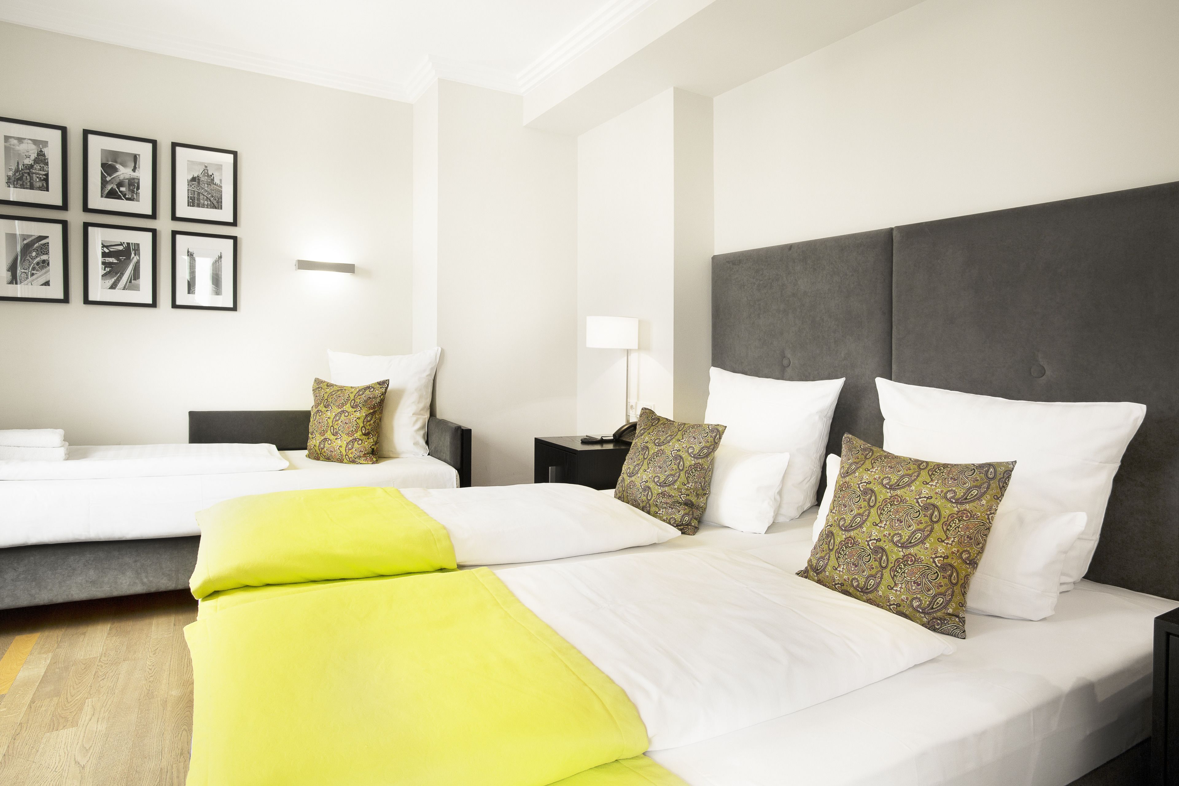 Een hotelkamer met twee bedden, een groot grijs hoofdeinde en een muur met zes zwart-witfoto's.