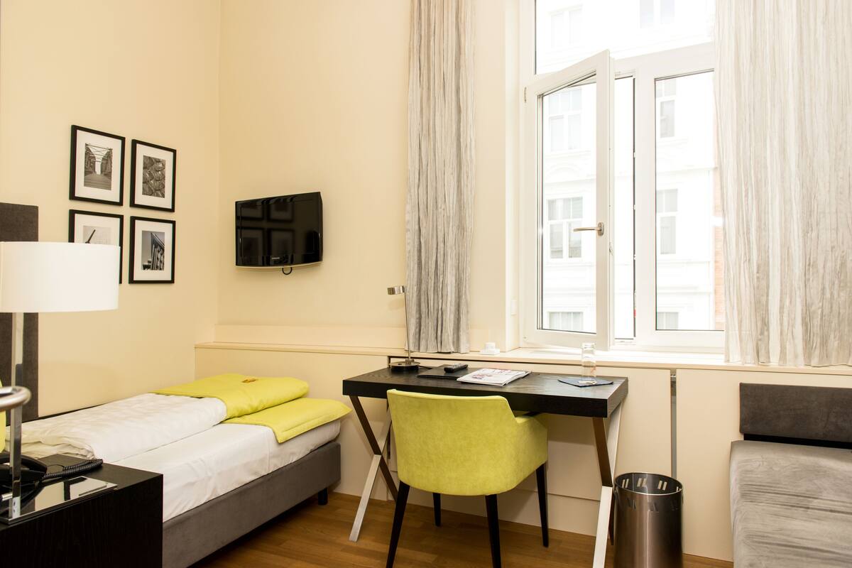 Quarto de hotel com cama, escrivaninha, cadeira, televisão e janela com cortinas.