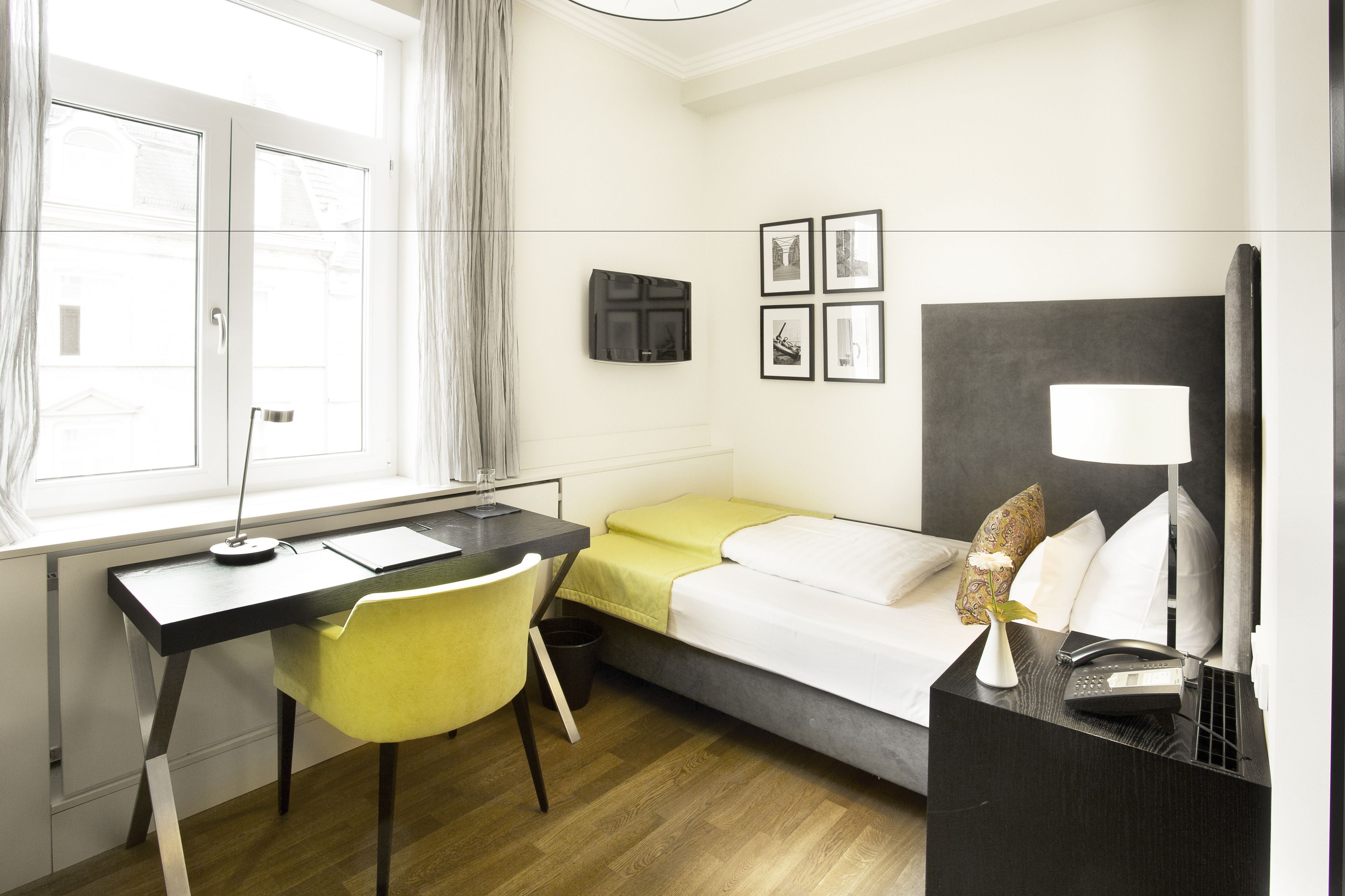 A compact hotel room with a desk, chair, bed, bedside table, lamp, and a small window with curtains.
