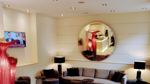 Un salon moderne avec un canapé d’angle, des poufs rouges, un miroir rond et un téléviseur à écran plat.