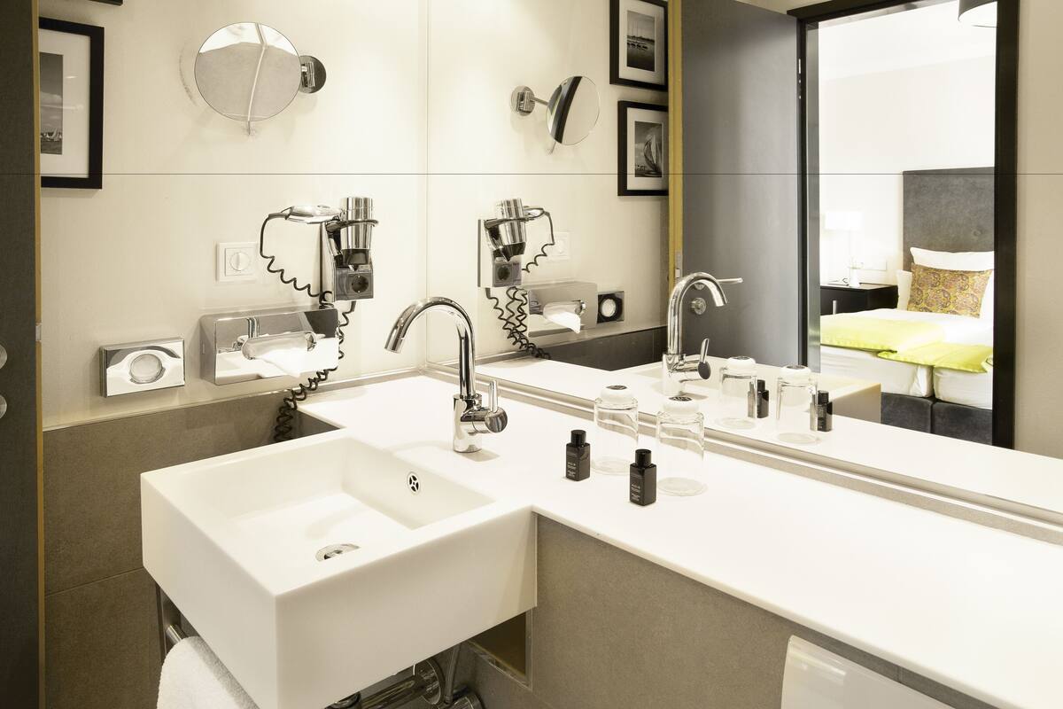 Casa de banho moderna com duas lavatórios, um espelho grande e vista para um quarto com cama e mesa de cabeceira.