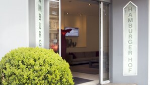 L’entrée d’un hôtel, avec une enseigne indiquant « Hamburger Hof » et une grande plante en pot au premier plan.