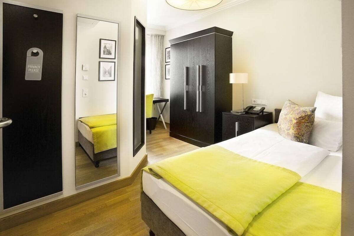 Quarto de hotel com cama, guarda-roupa, secretária com candeeiro e espelho.