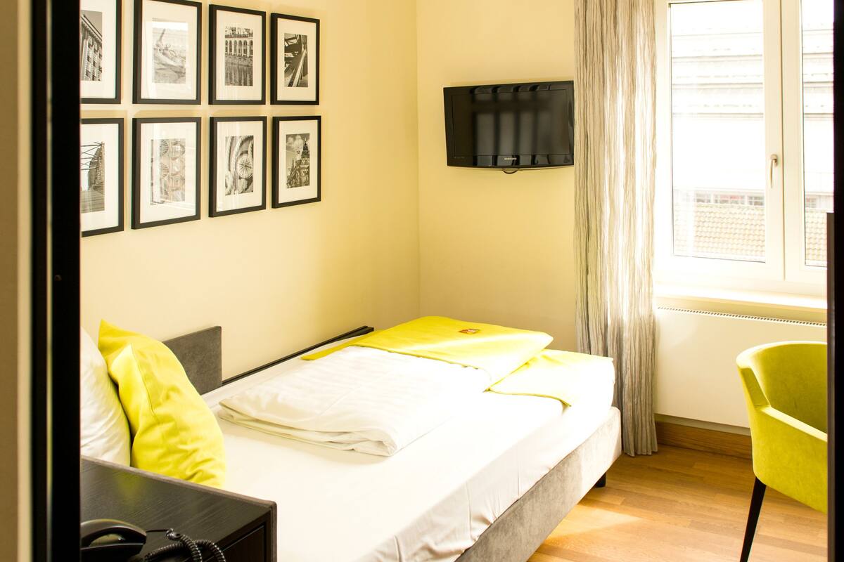 Quarto de hotel com cama, escrivaninha com telefone, poltrona, televisão e quadros nas paredes.