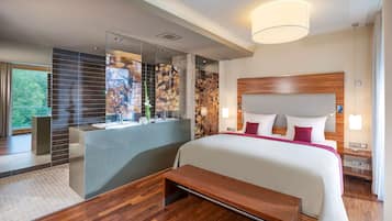 A modern hotel room with a large bed, a wooden headboard, a bench, a bathroom with a glass partition, and a large window offering a view of trees.