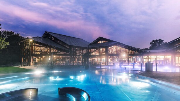 A modern hotel with a large outdoor swimming pool, covered seating areas, and a well-lit entrance.