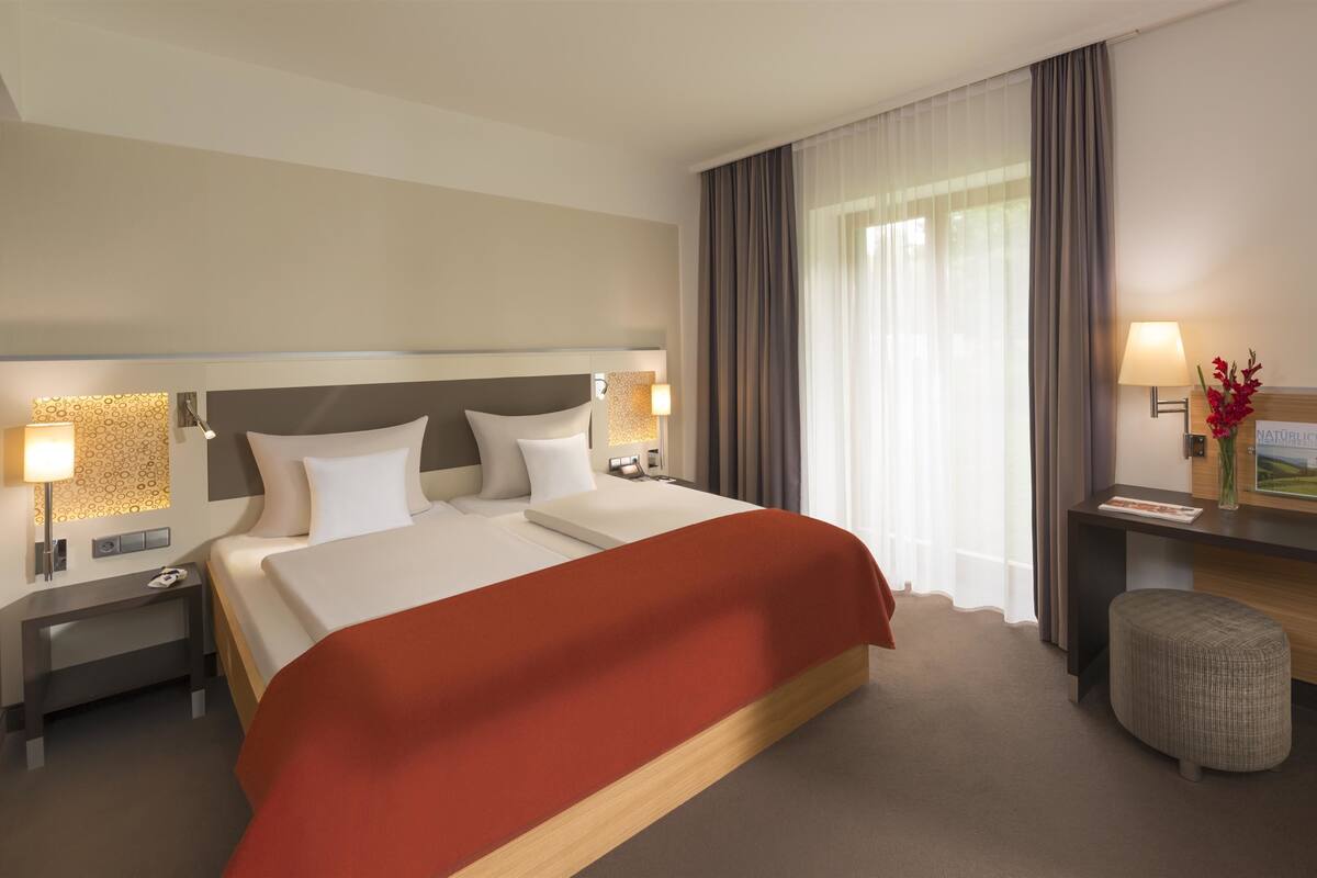 Habitació d'hotel amb un llit, làmpades de nit, una televisió, un escriptori amb una làmpada i un petit tamboret.