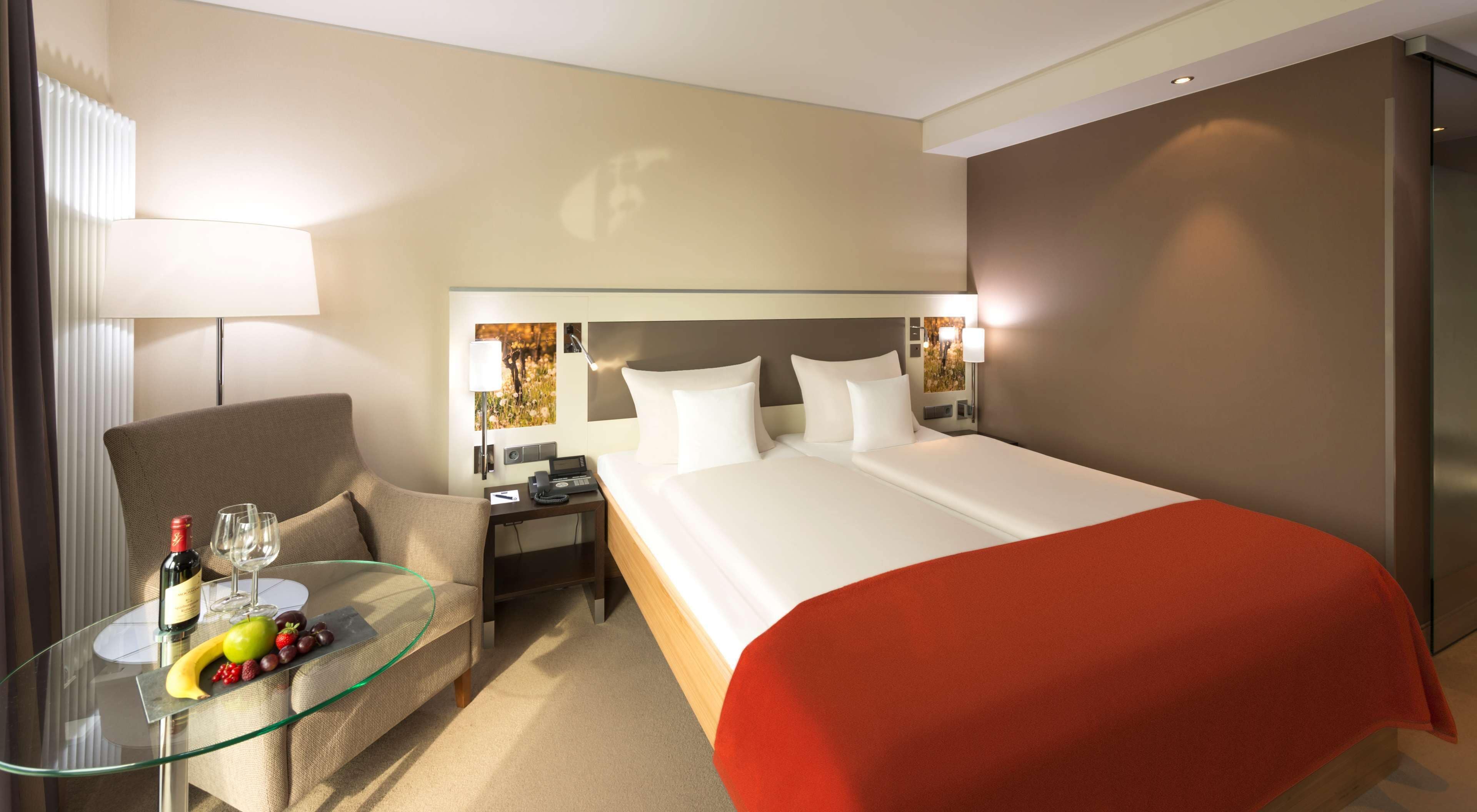 A hotel room with a bed, a glass table with fruit and a bottle, a chair, and a desk with a lamp.