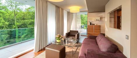 A modern living room with a glass coffee table, a brown leather ottoman, a purple sofa, and a view of a forest through large windows.