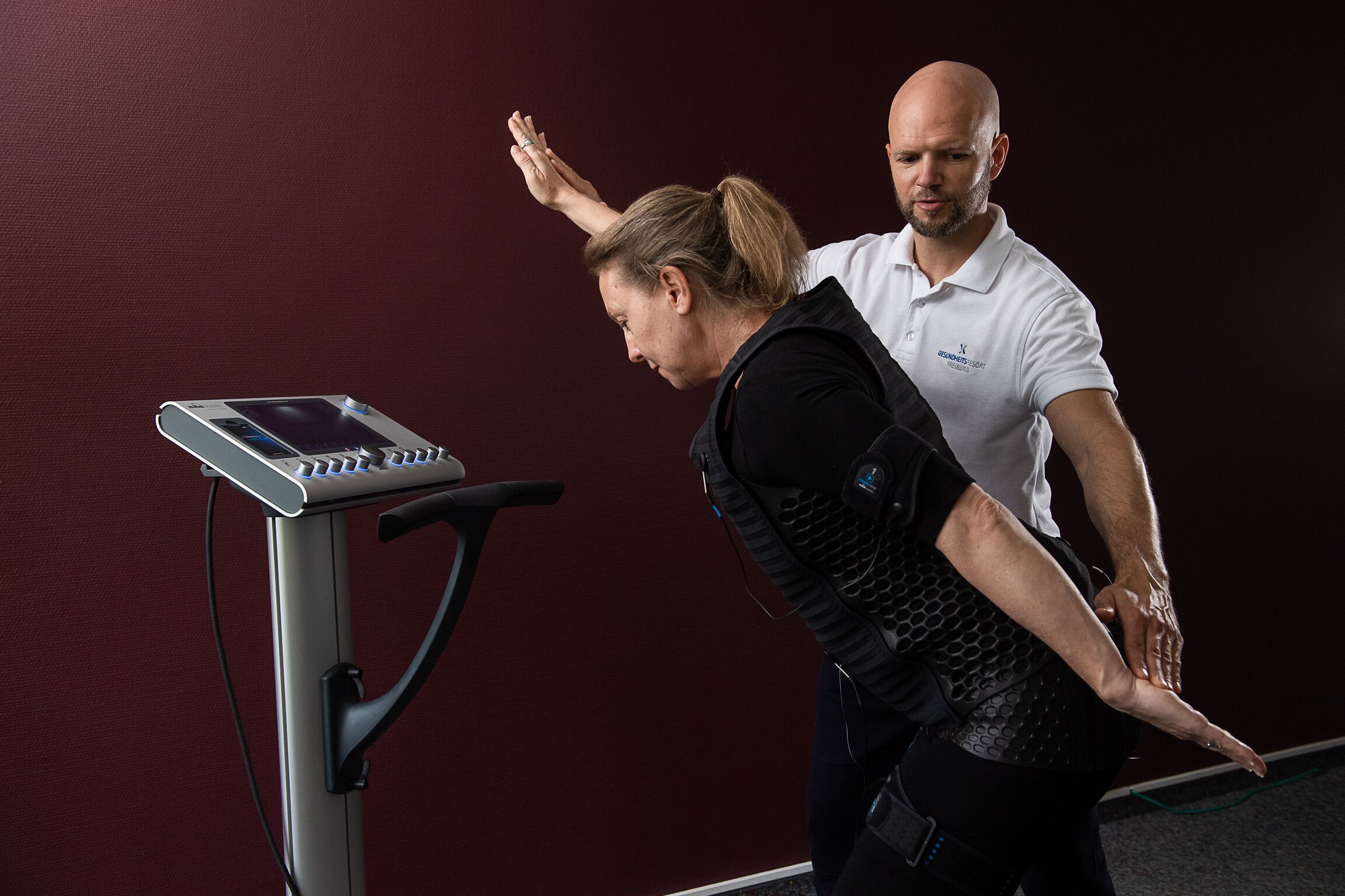 Eine Frau in einer schwarzen Weste führt eine Physiotherapie-Übung auf einem stationären Fahrrad mit Digitalanzeige durch, unterstützt von einem Mann in einem weißen Poloshirt.