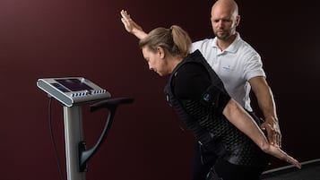 Eine Frau in einer schwarzen Weste führt eine Physiotherapie-Übung auf einem stationären Fahrrad mit Digitalanzeige durch, unterstützt von einem Mann in einem weißen Poloshirt.