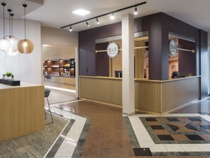 A modern hotel reception area with a wooden reception desk, a B&B sign, and a view into the hotel's interior.
