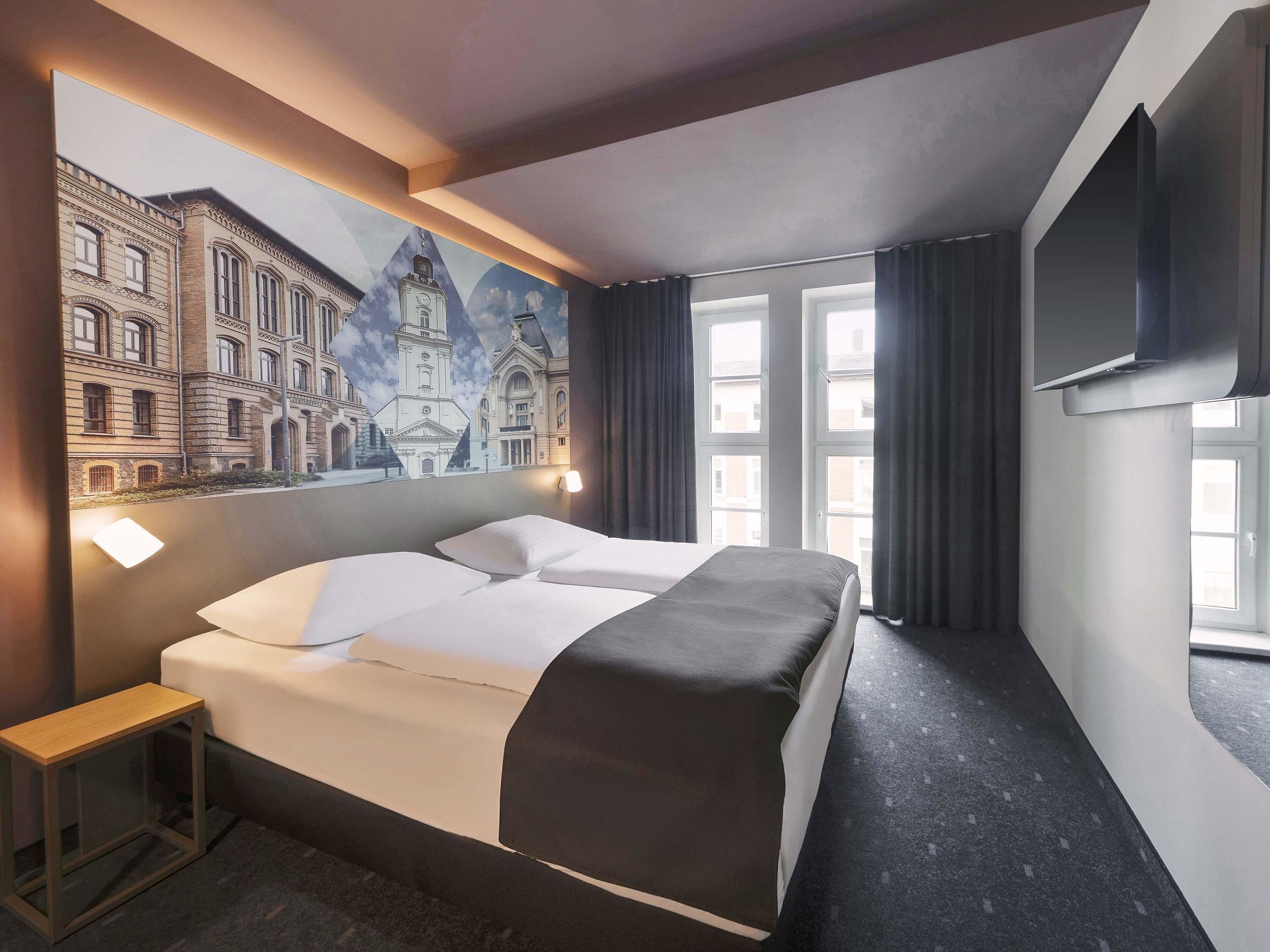 A hotel room with a bed, a small table, a TV mounted on the wall, and a large window with curtains.