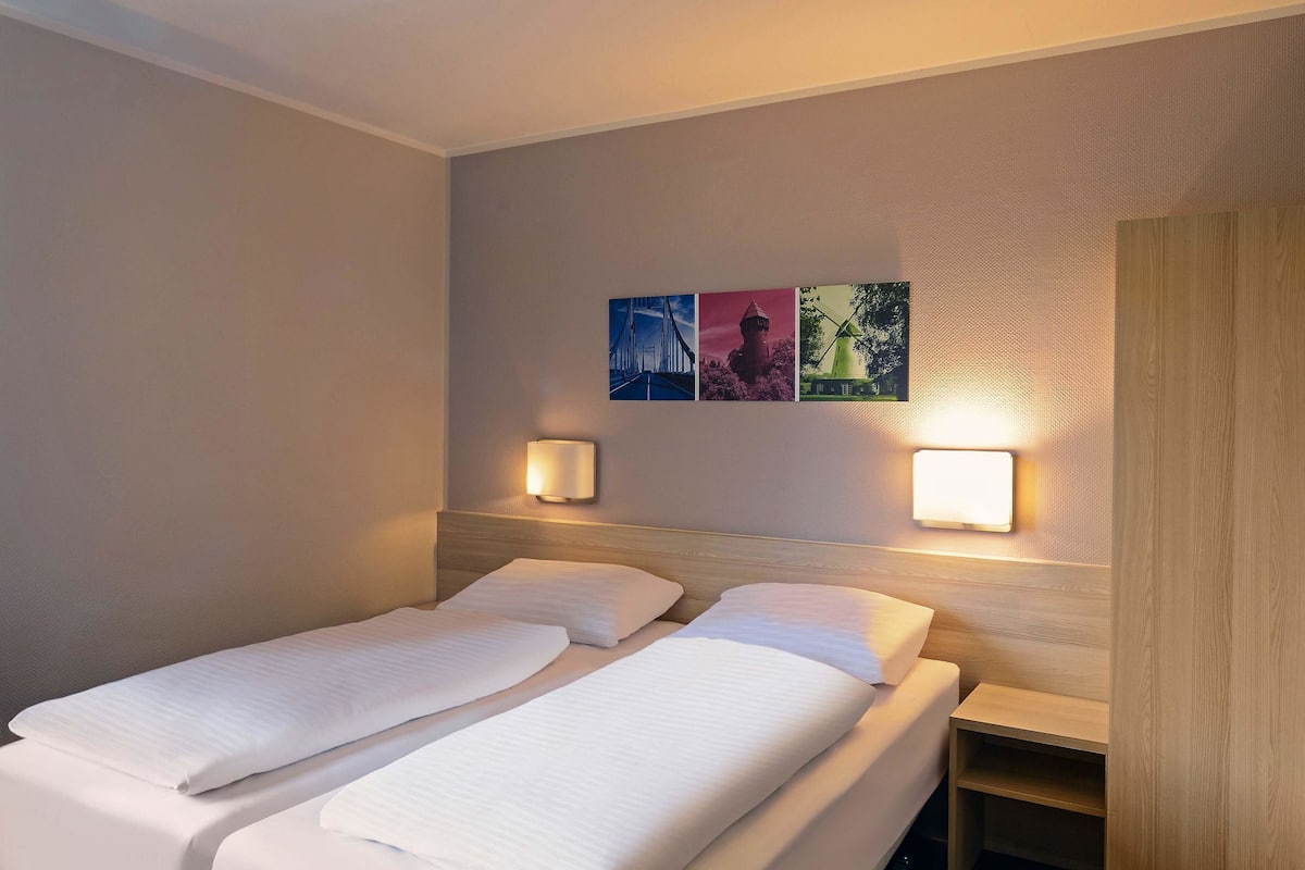 Habitación de hotel con dos camas, una mesita de noche y lámparas fijadas a la pared.
