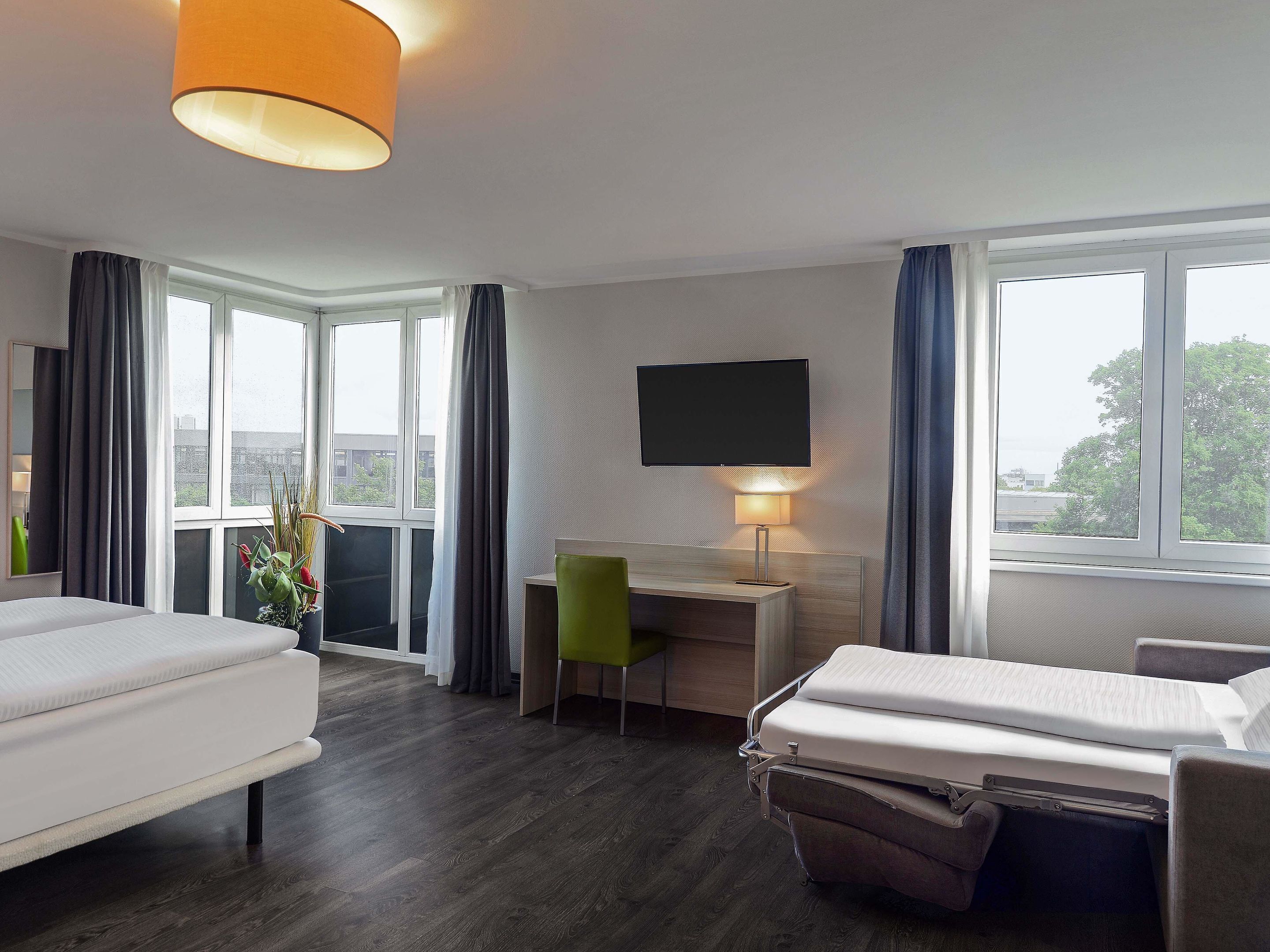 A hotel room with a bed, desk, chair, TV, and large windows.