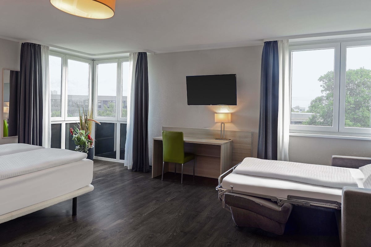 Una habitación de hotel con cama, escritorio, silla, televisor y ventanas amplias.