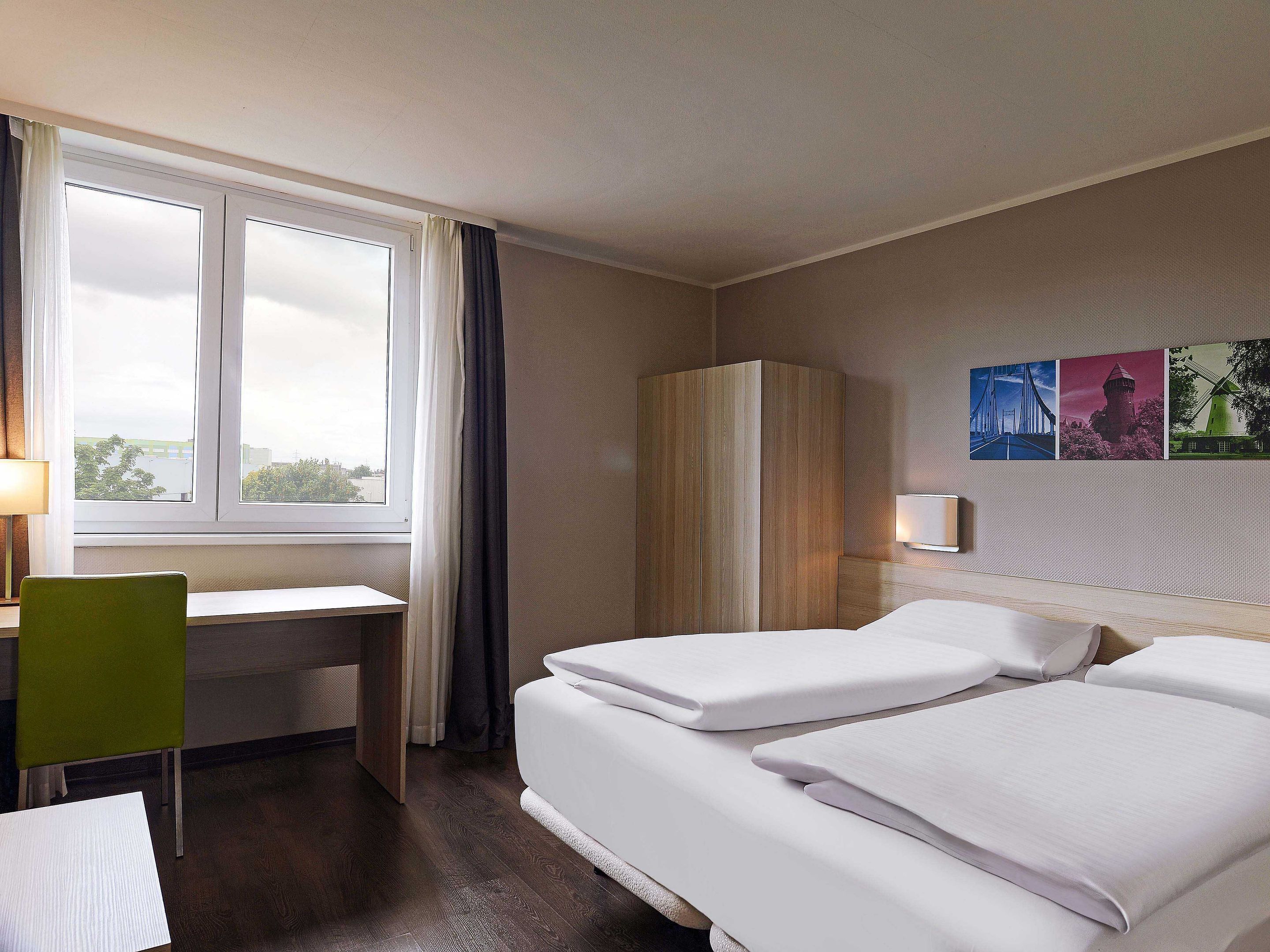 A hotel room with a desk, two beds, a wardrobe, a window with curtains, and wall art.