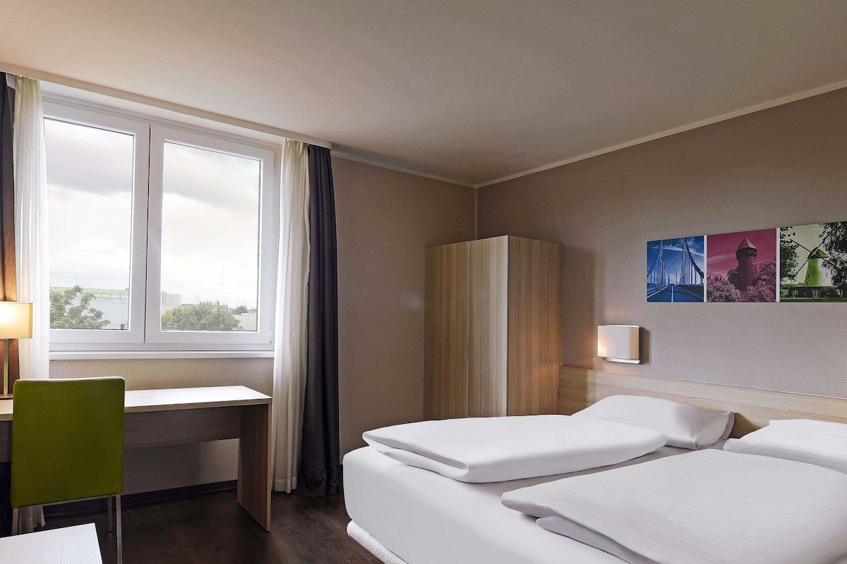 Habitación de hotel con escritorio, dos camas, armario, ventana con cortinas y decoración en la pared.