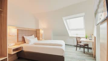 A hotel room with a bed, bedside table, chair, desk, and a window.