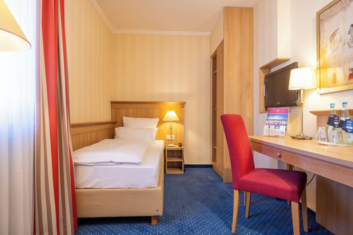A hotel room with a bed, a desk, a chair, a TV, and a lamp.