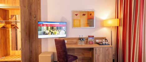 A hotel room with a wooden desk, a red velvet chair, a television mounted on the wall, a painting, and a lamp.
