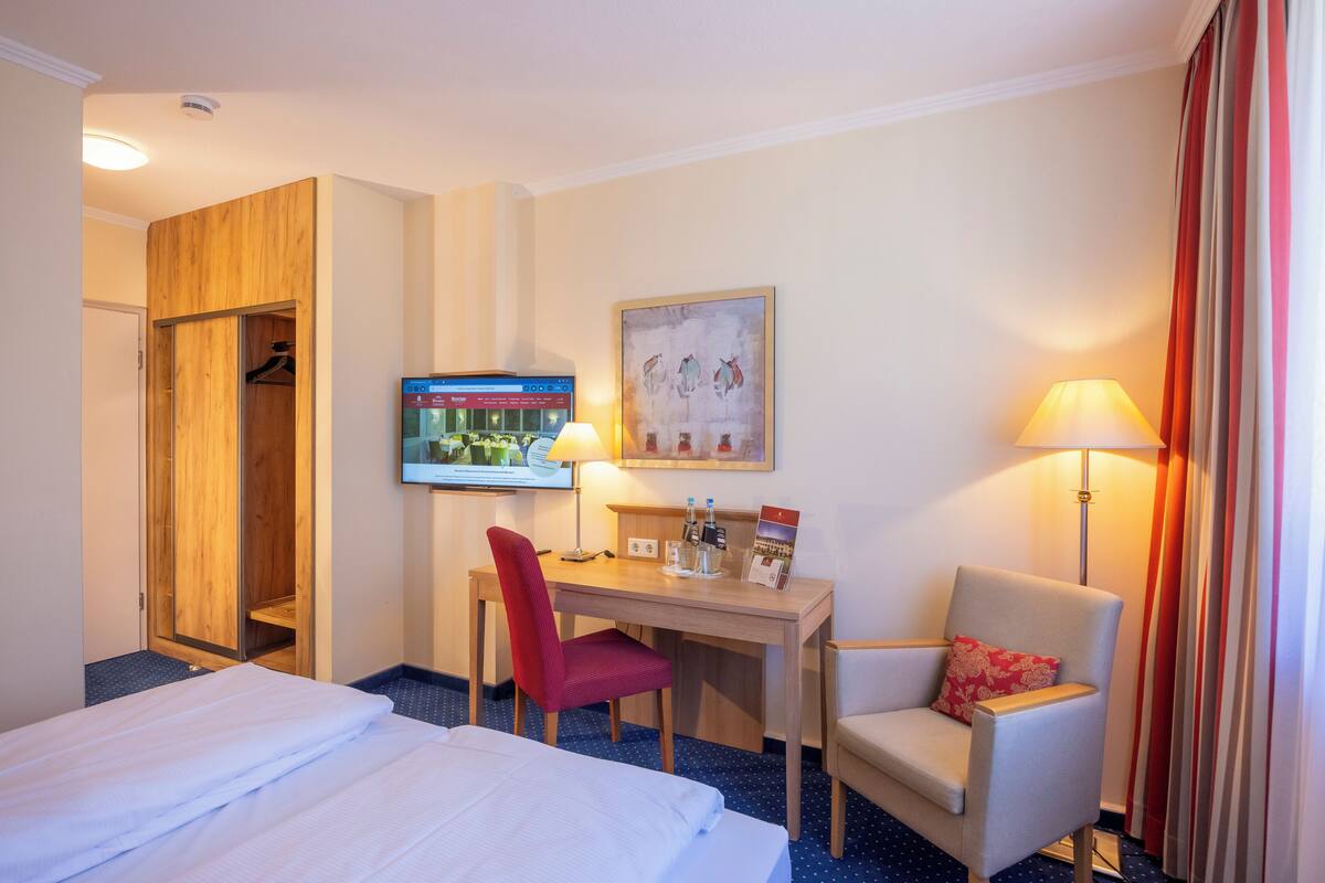 A hotel room with a bed, a desk with a chair, a TV, a painting, and a lamp.