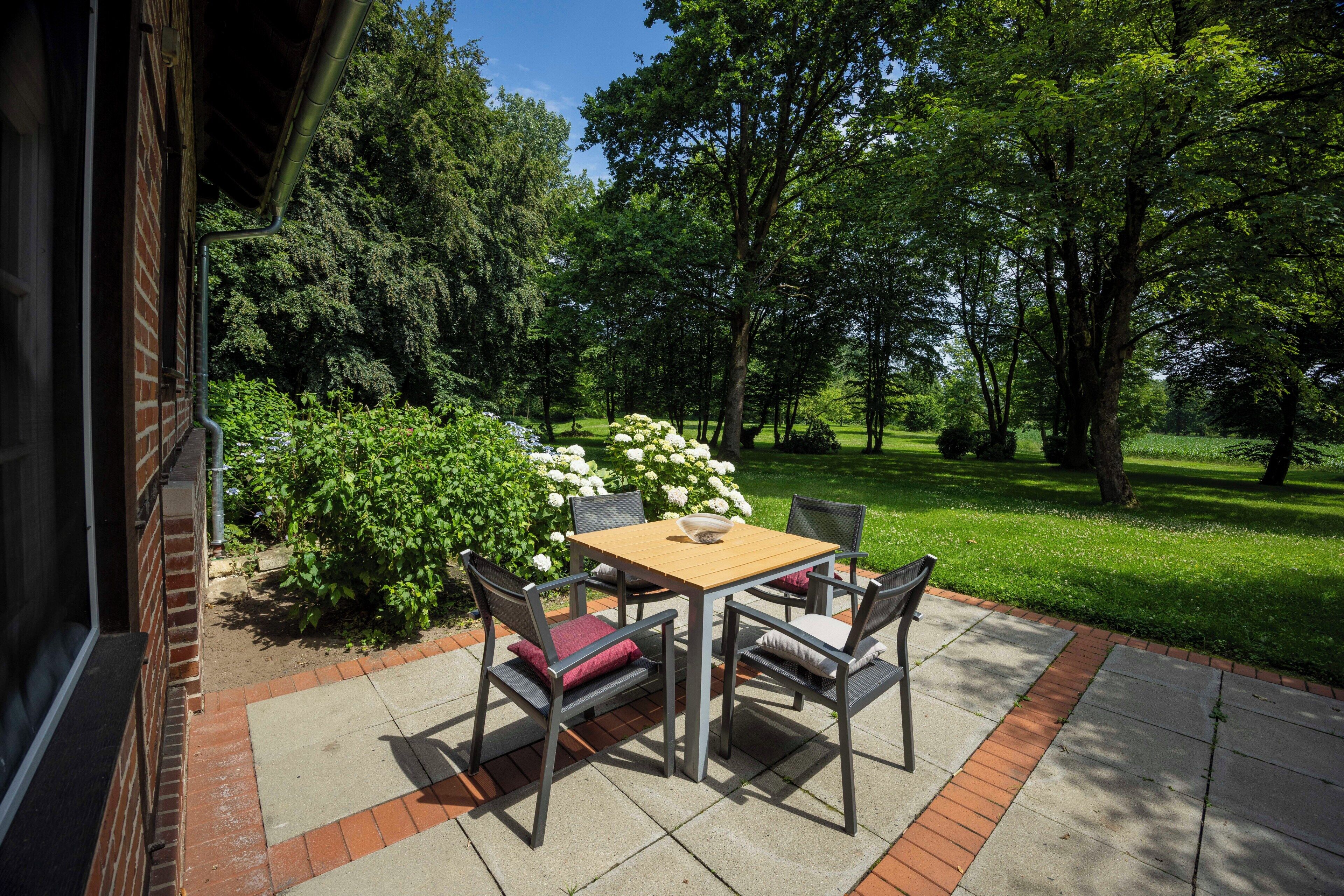 Eine Terrasse im Freien mit Tisch und Stühlen, umgeben von viel Grün und Bäumen.
