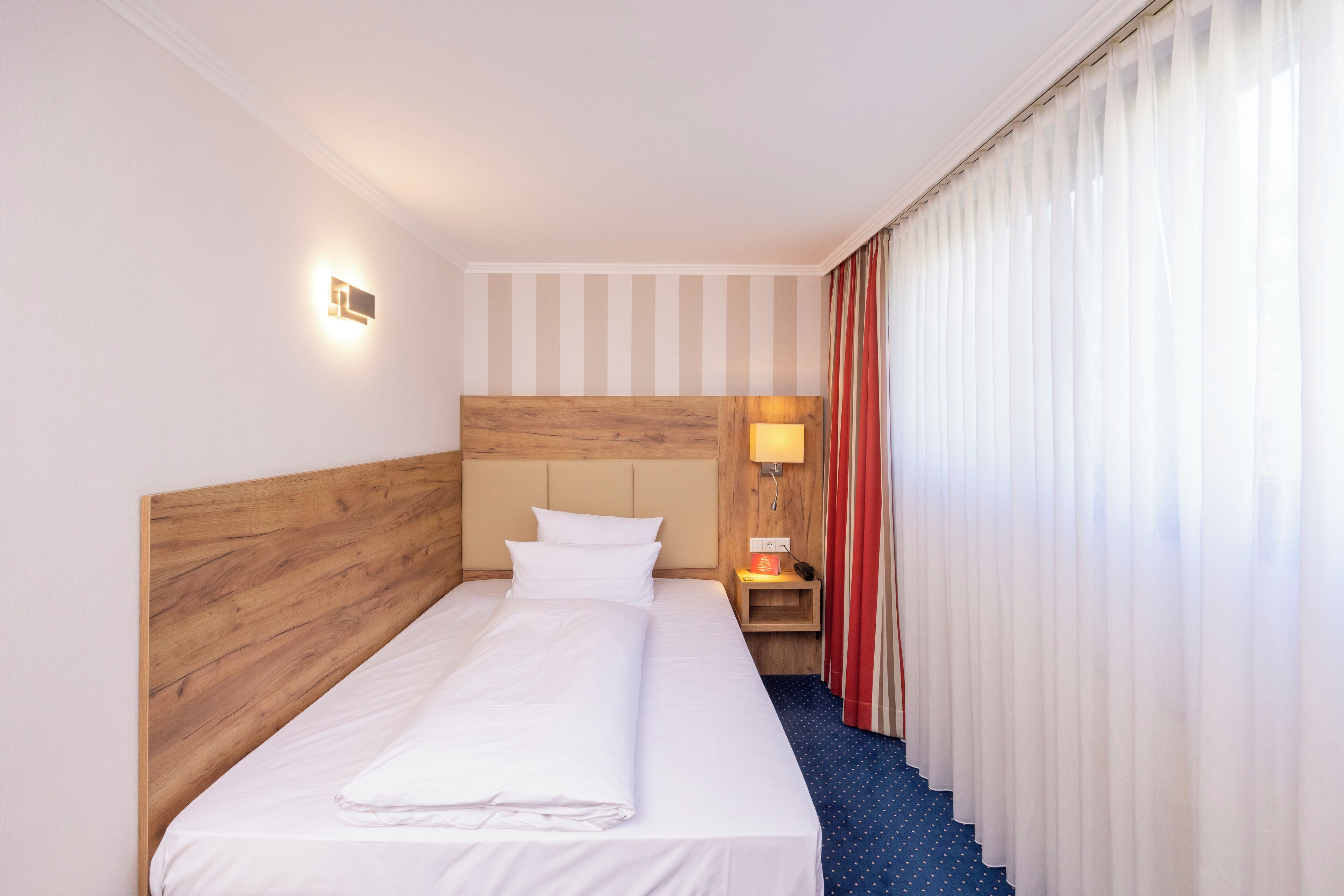 Habitación de hotel con cabecera de madera, cama bien tendida, mesita de noche con lámpara y ventana con cortinas translúcidas.