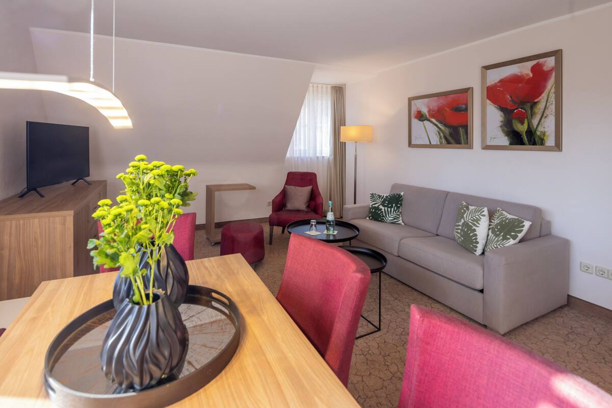 A modern living room with a wooden dining table, red chairs, a grey sofa, and a TV on a stand.