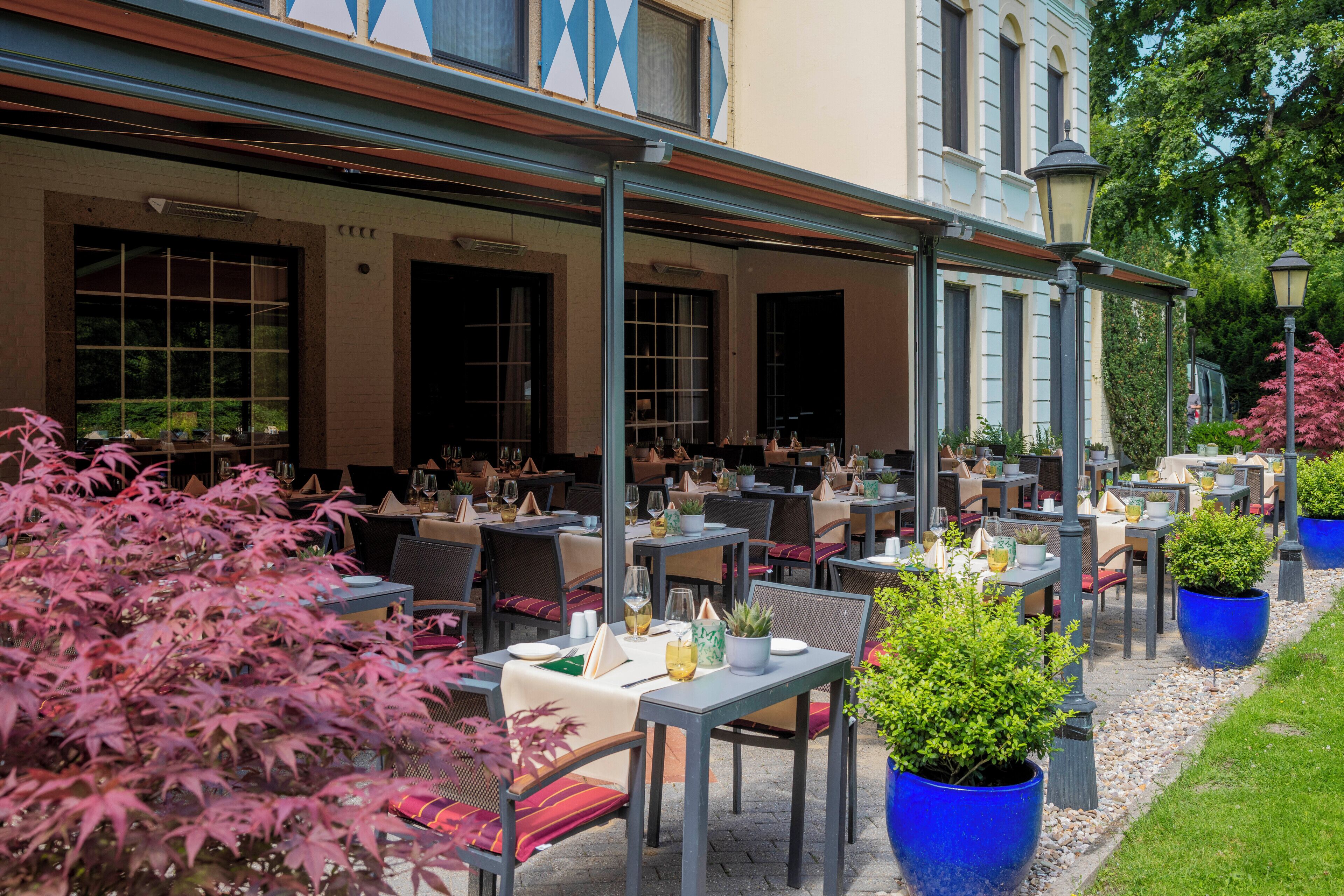 An outdoor dining area with multiple tables and chairs, surrounded by potted plants and a building with large windows.