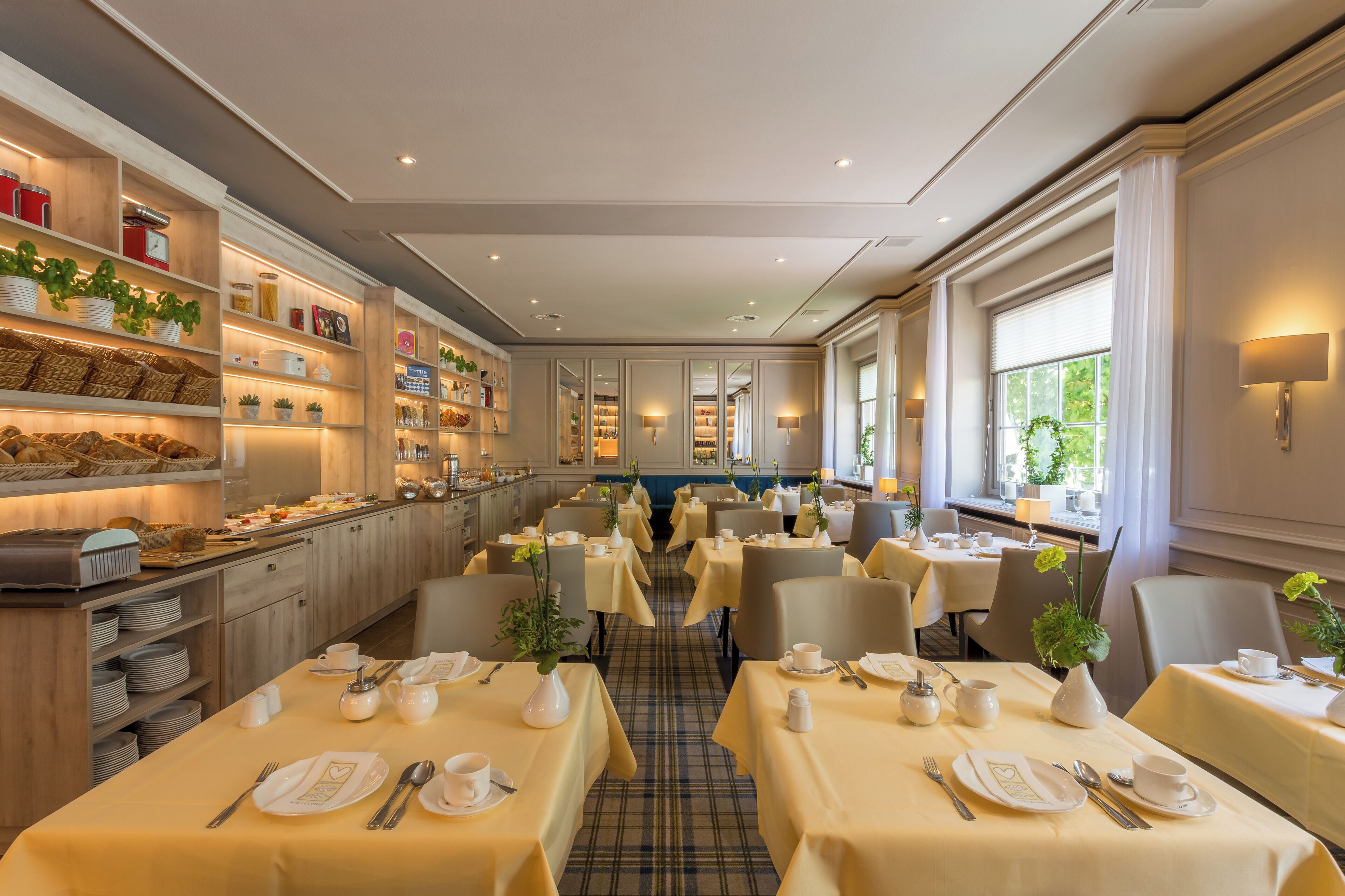 Comedor de hotel con varias mesas dispuestas para el desayuno, que incluye un bufé con diversas opciones de comida y una selección de plantas.