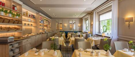 Comedor de hotel con varias mesas dispuestas para el desayuno, que incluye un bufé con diversas opciones de comida y una selección de plantas.
