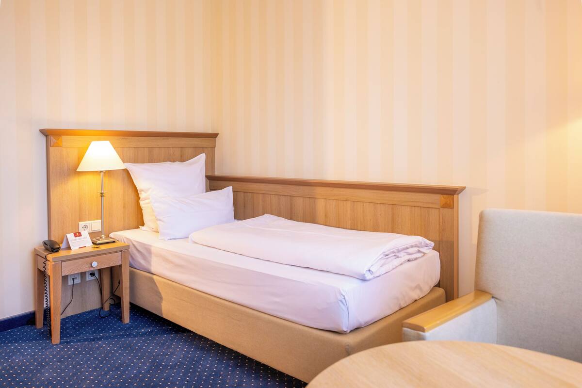 A hotel room with a wooden headboard, a single bed with white linens, a bedside table with a lamp, and a chair.
