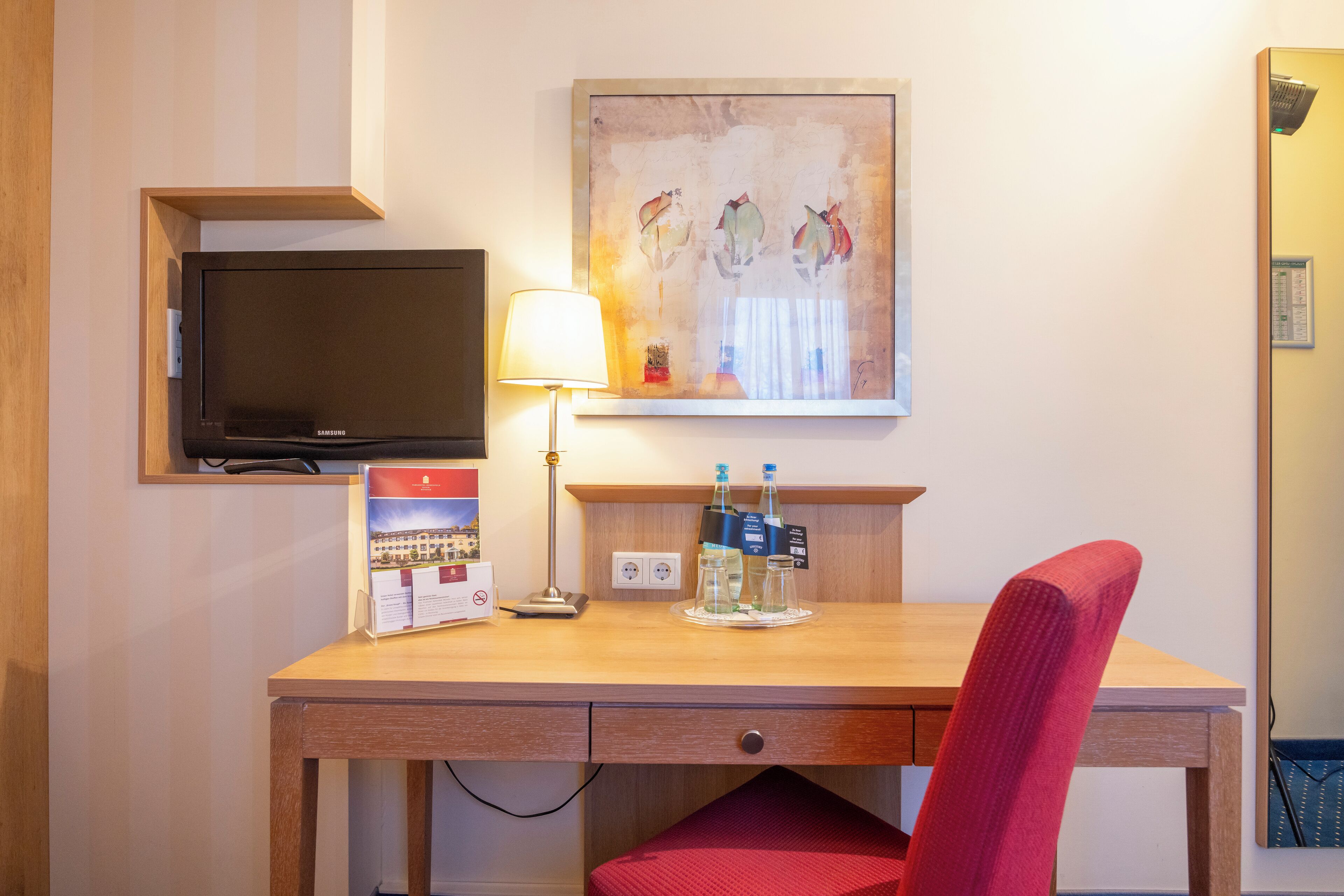 A hotel room with a wooden desk, a television, a lamp, a painting, and a red chair.