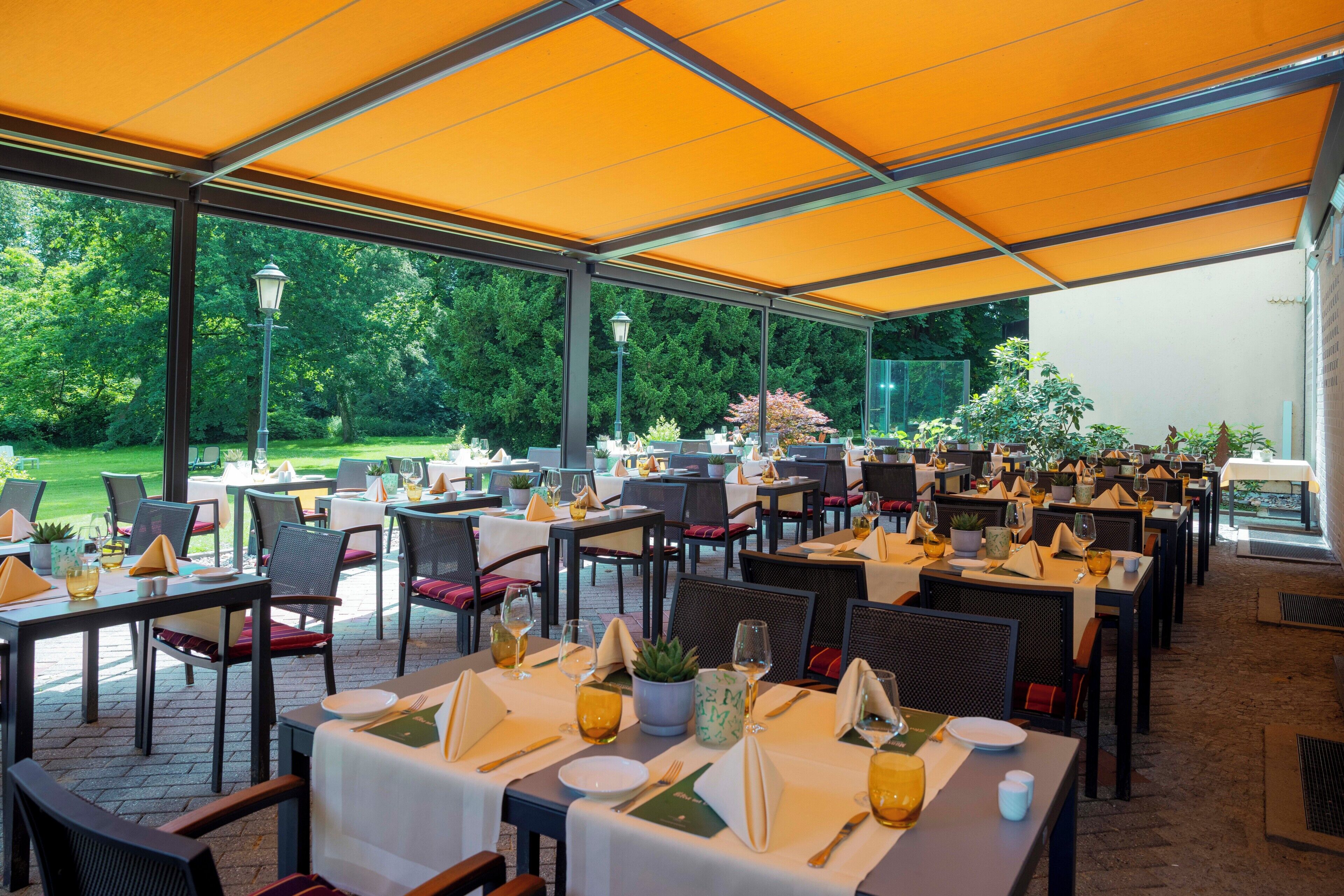 An outdoor restaurant with tables set for dining, covered by a yellow canopy, and surrounded by greenery.