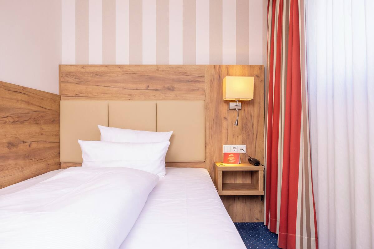 A hotel room with a wooden headboard, a bedside table with a lamp, and a window with striped curtains.