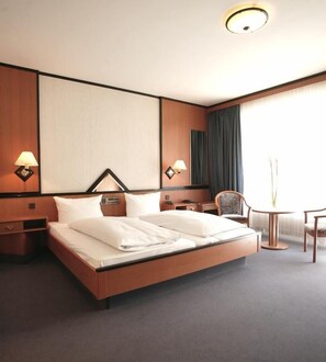 A hotel room with a large bed, bedside lamps, a chair, a small table, and a window with curtains. - Top Hotel Senator Munich (Munich)