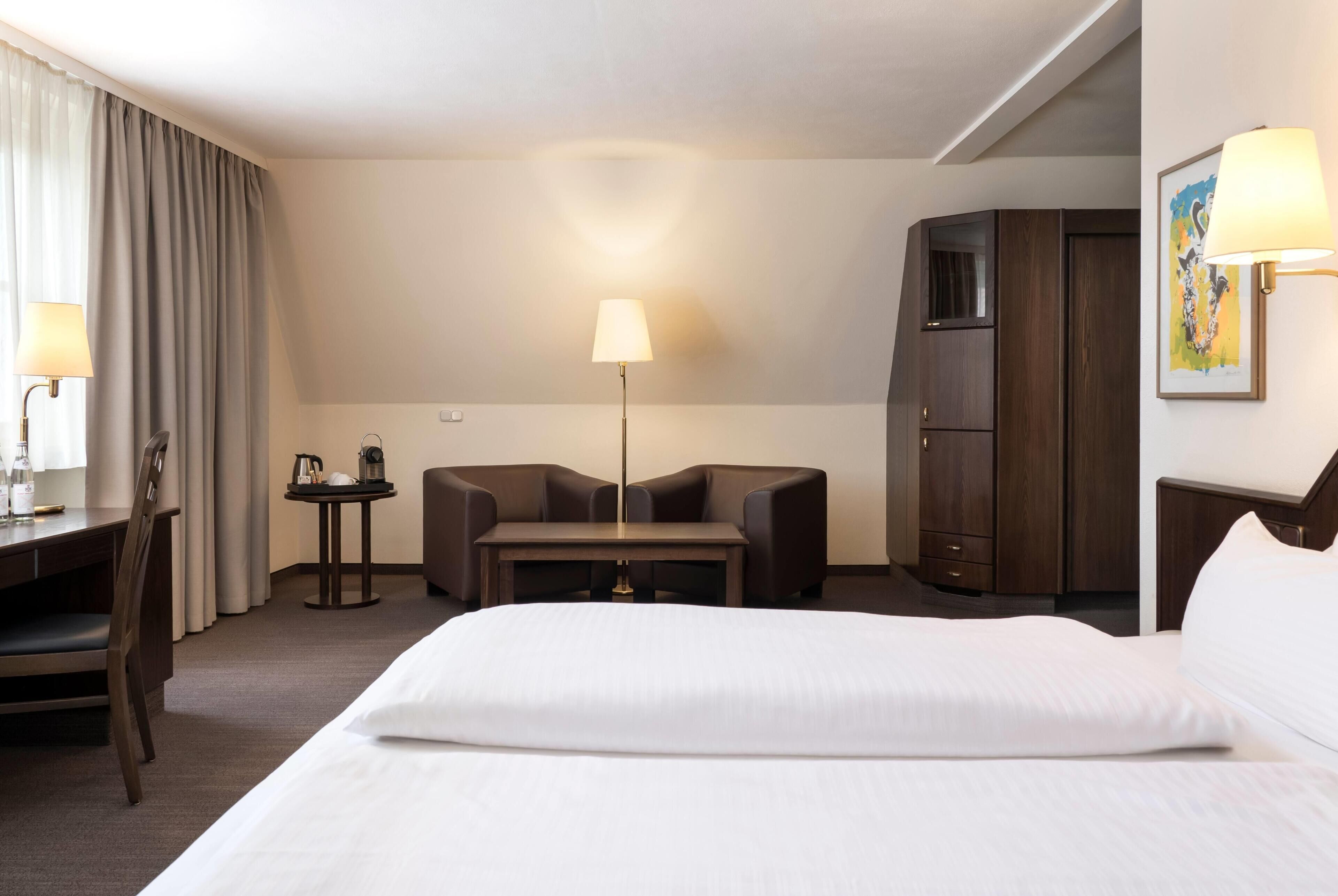A hotel room with a bed, a desk, a chair, a table, a lamp, a wardrobe, and a painting on the wall.