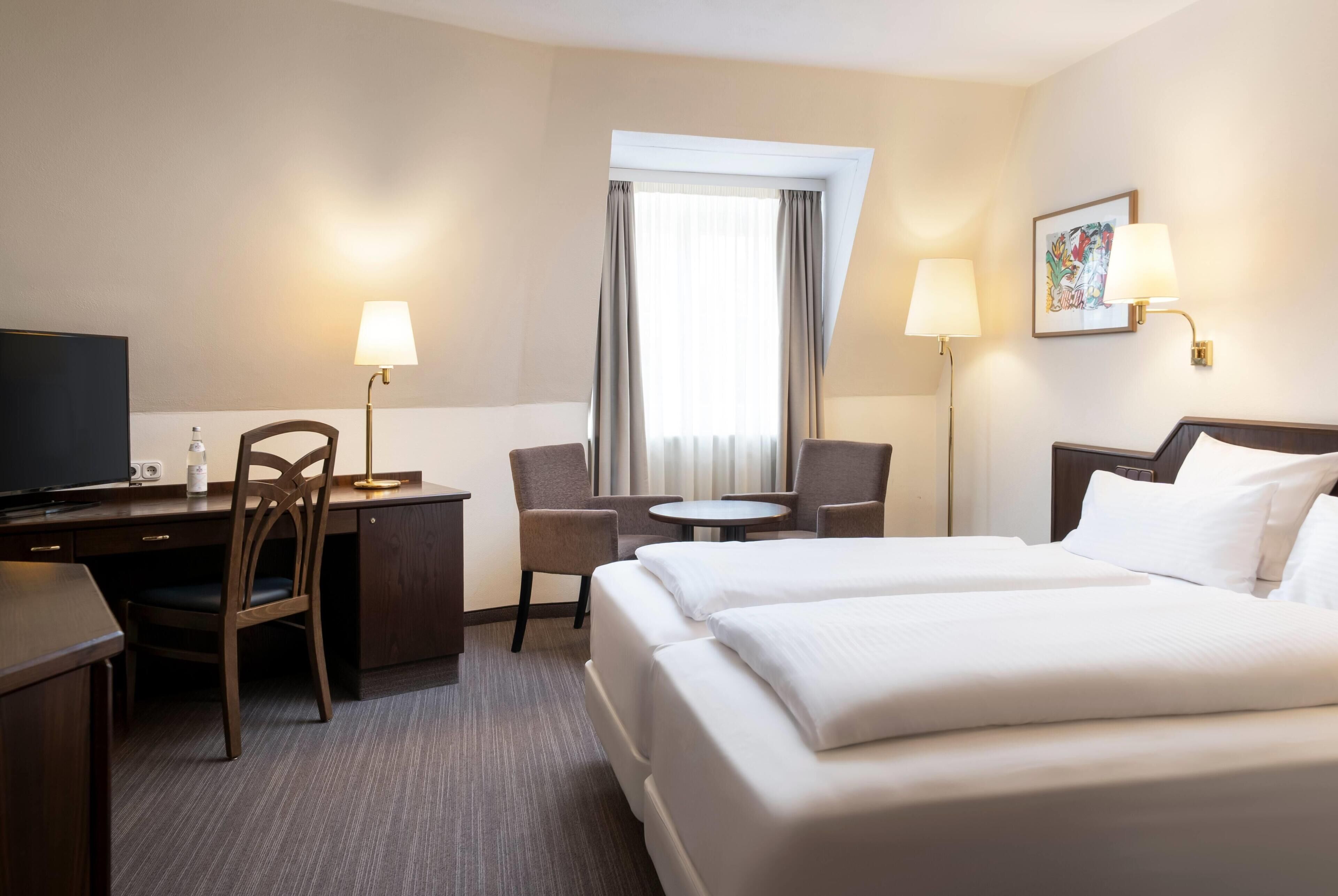 A hotel room with a bed, a desk, a chair, a small table, and two lamps.