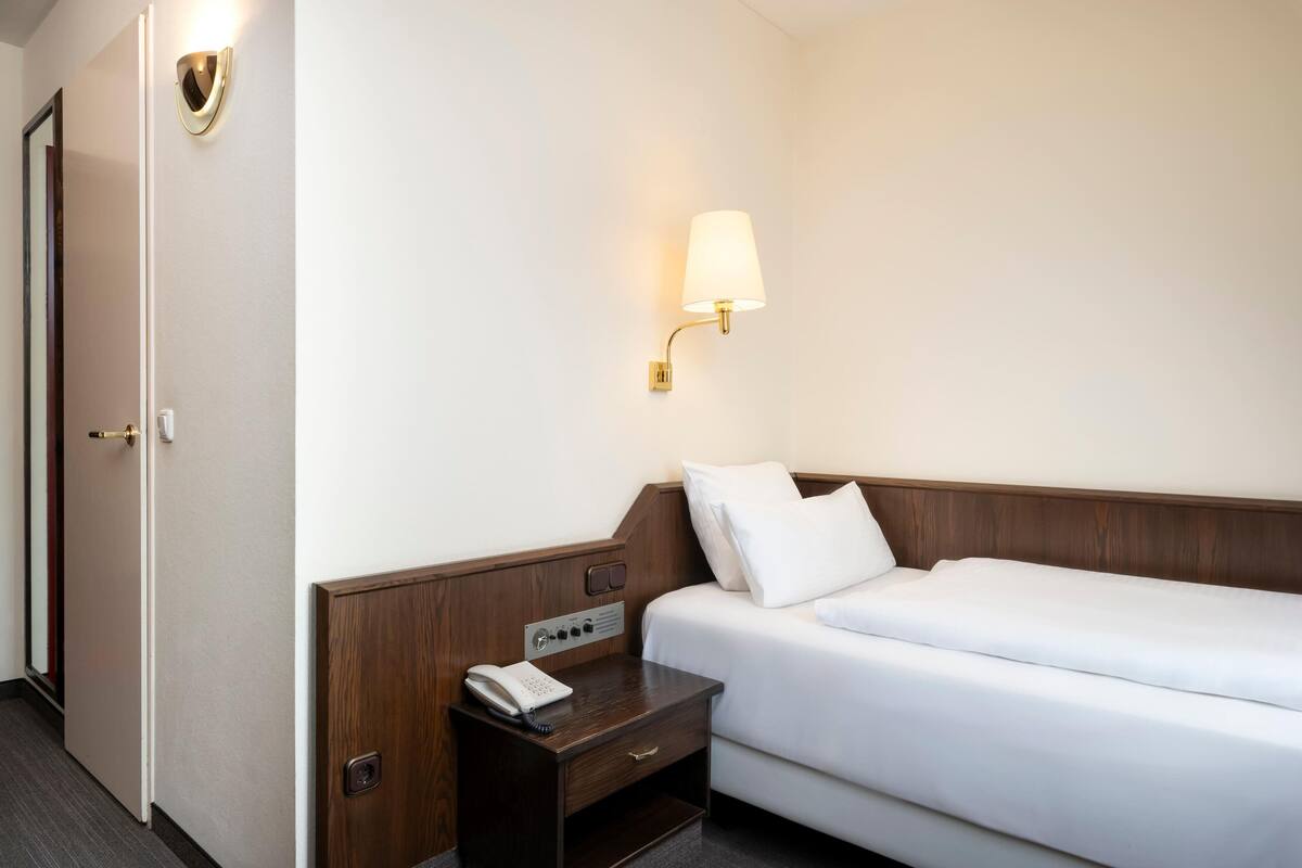 Habitación de hotel con una cama individual, una mesita de noche con un teléfono, una lámpara de pared y una puerta.