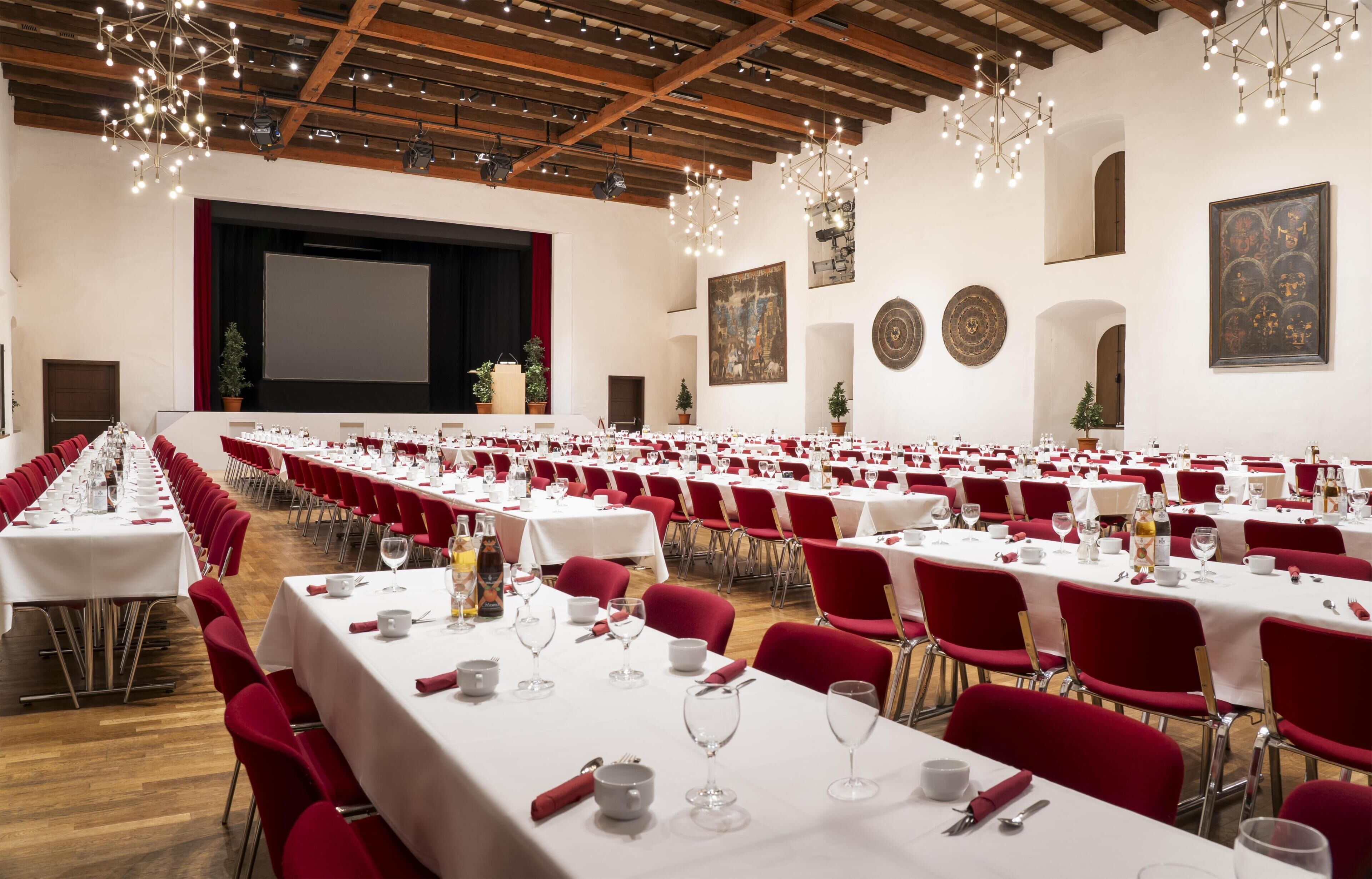 A large conference room with rows of tables set for dining, red chairs, and a stage with a screen.
