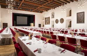 Een grote congreszaal met rijen tafels gedekt voor een maaltijd, rode stoelen en een podium met een scherm.