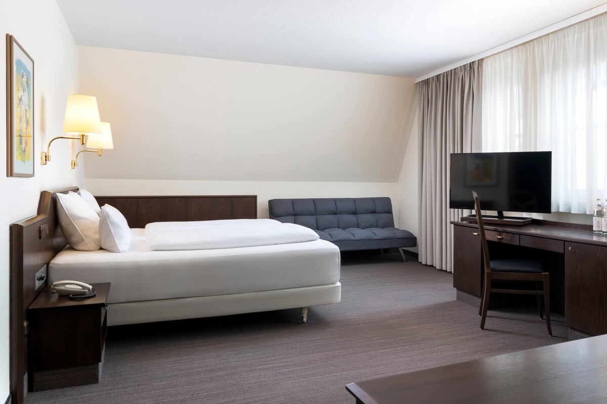 A hotel room with a bed, a desk with a television, a chair, and a lamp.